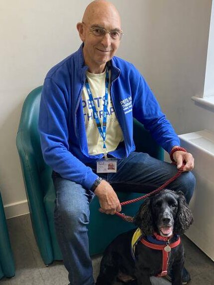 Mike Tev wearing a blue jacket, yellow t-shirt, jeans and NHS lanyard. Holding Patch, the therapy dogs lead. Both are looking at the camera.