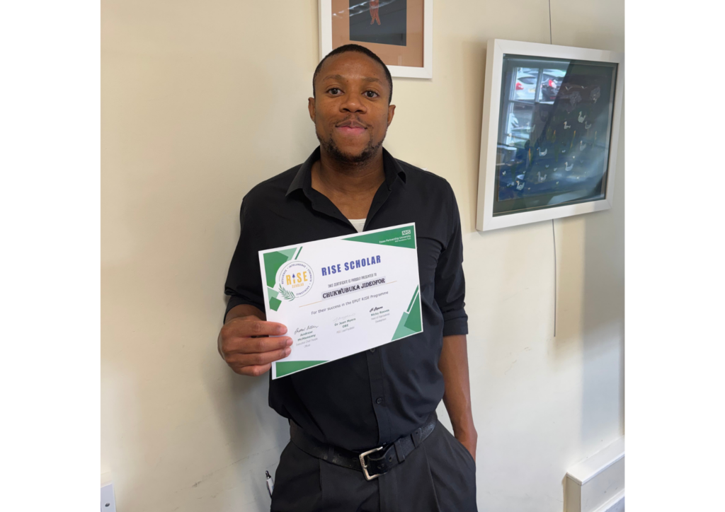 Chukwuebuka Jideofor standing in front of a light coloured wall proudly holding his RISE certificate