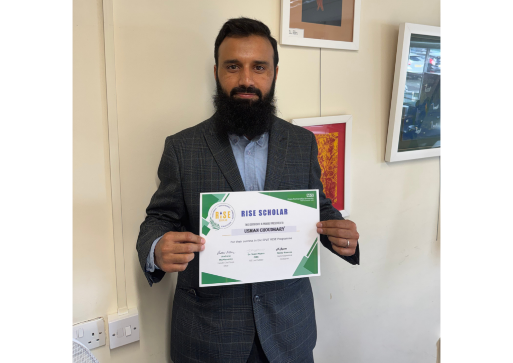 Usman Choudhary standing in front of a light coloured wall proudly holding his RISE certificate