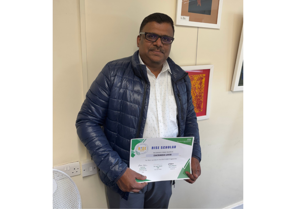 Zacharia Louis standing in front of a light coloured wall proudly holding his RISE certificate