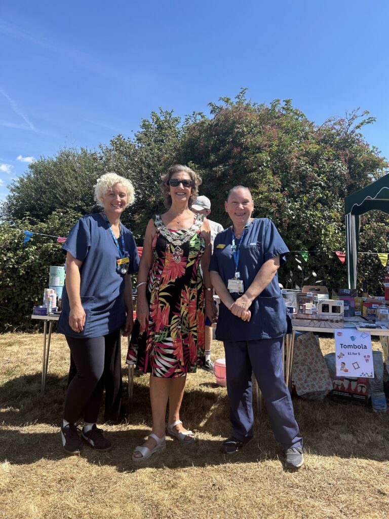 Three women smiling together outdoors at a community event, standing in front of stalls under a clear blue sky. Two wear dark blue healthcare scrubs and lanyards, while the woman in the center wears a colorful tropical print dress and a mayoral chain.