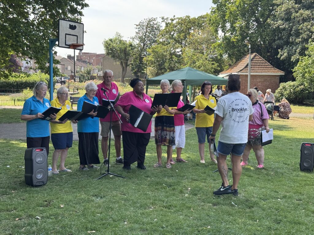 A diverse outdoor choir wearing colorful polo shirts sings together in a grassy park, holding music folders as a choir director conducts them