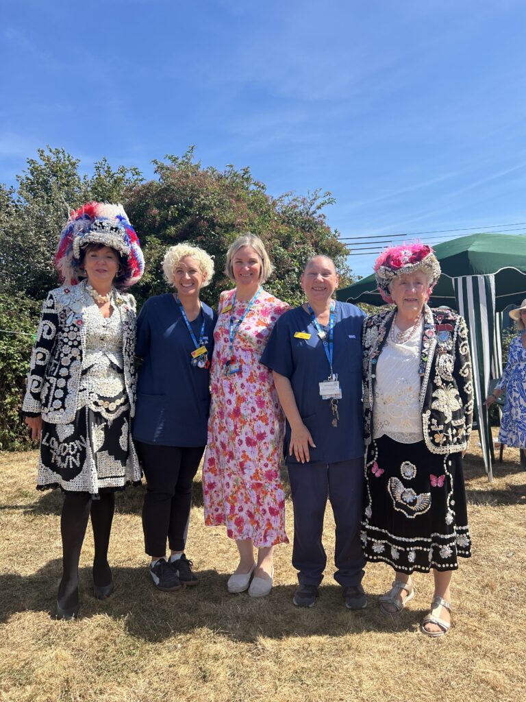Five smiling women standing together outside on a bright, sunny day. Two women on the ends are dressed in full Pearly Queen traditional outfits with ornate mother-of-pearl buttons and feathered hats, flanked around three care home staff members.
