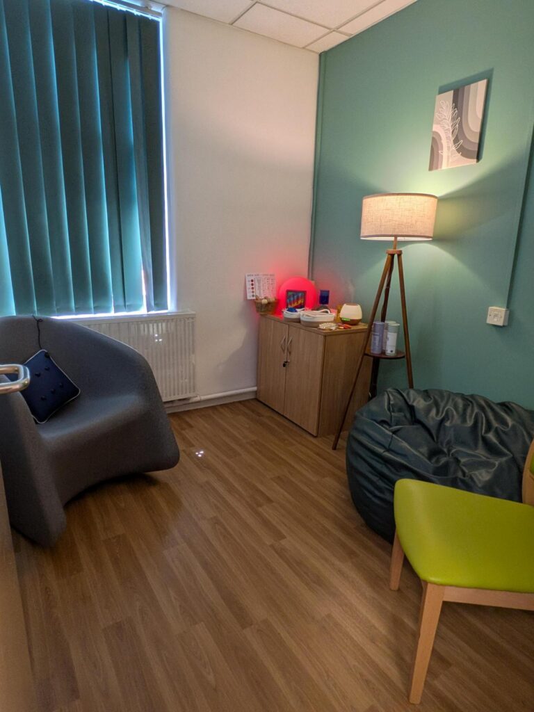 A peaceful therapeutic room corner featuring a grey curved armchair, a large blue beanbag, and a small lime green chair. In the background, a wooden cabinet holds sensory tools and a warm lamp provides a soft, calming light against a teal wall.