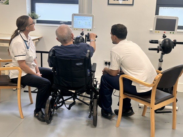 A patient in a wheelchair using specialized digital rehabilitation equipment with support from two healthcare staff members at St Margaret's Hospital.