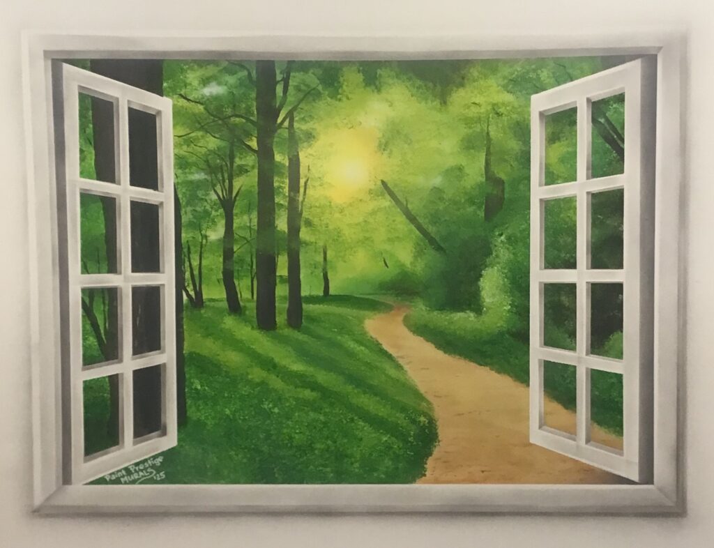 A mural painting depicting an open white window looking out into a serene, sunlit forest. A dirt path winds through lush green grass and tall trees toward a bright glowing sun in the background. The white multi-paned window shutters swing outward, creating a 3D illusion on the wall. In the bottom-left corner of the window frame, a small signature reads "Paint Prestige Murals '25"