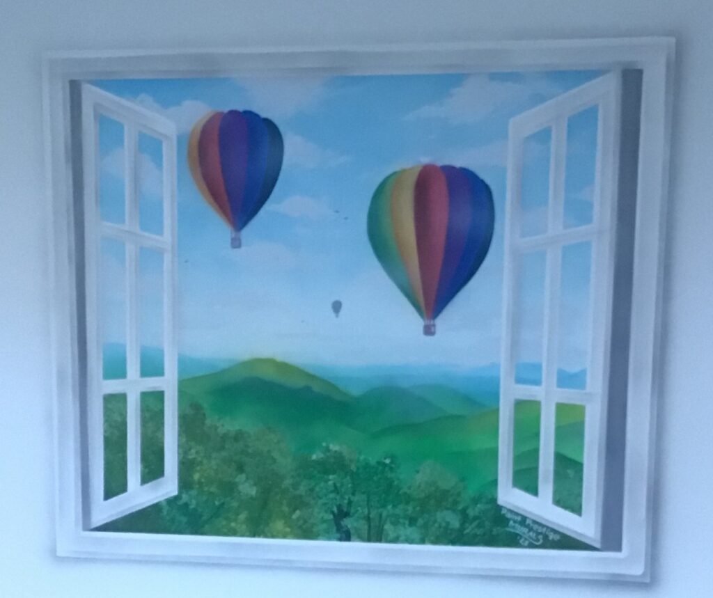 A wall mural painted to look like an open white window viewing colorful hot air balloons floating over green rolling hills and trees under a clear blue sky.