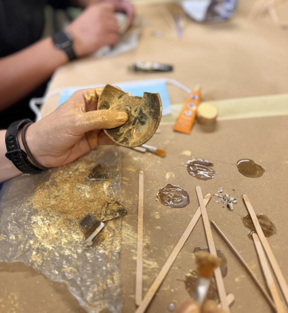 A small, broken Kintsugi bowl is being held up off a table by one hand. The Kintsugi and hand are covered in gold glitter.
There is also a few long, wooden lolly sticks on the table, surrounded by dabs of glue and glitter.