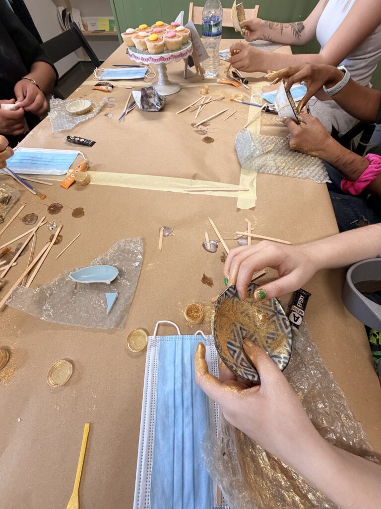 A small blue and white patterned Kintsugi bowl is covered in gold glitter and being held up off a table by two hands. The image also shows the full length of the table, and a few other peoples hands, dabs of glue and long lolly sticks.