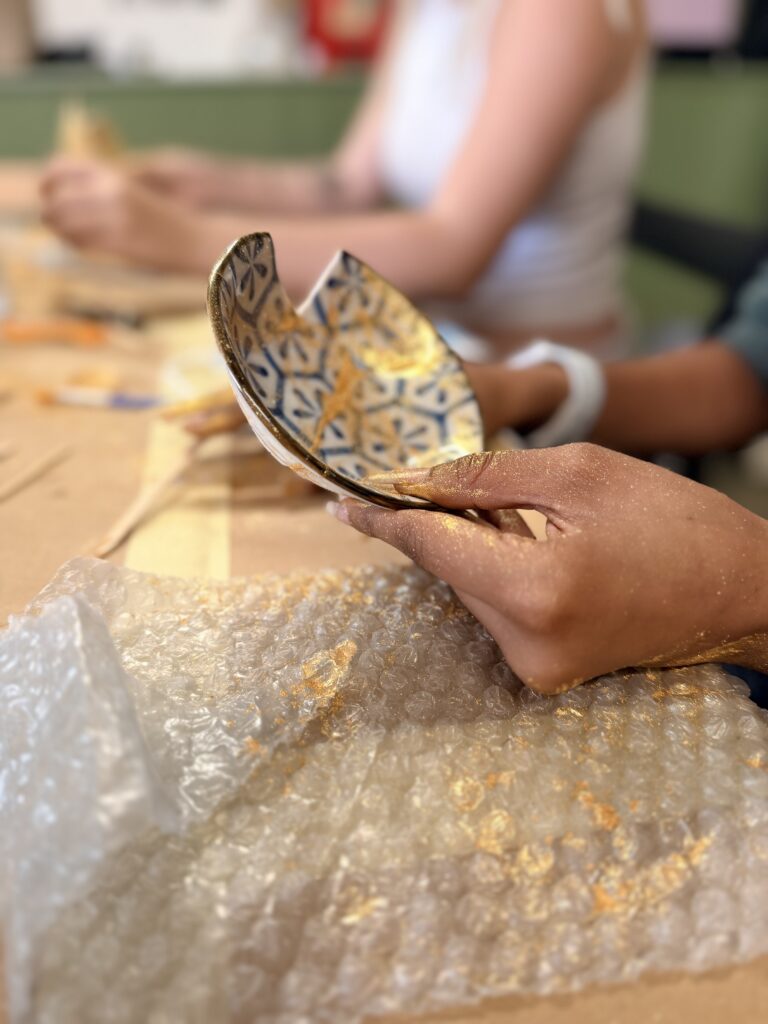 A small, broken Kintsugi bowl is being held up off a table by one hand. the Kintsugi is covered with gold glitter..