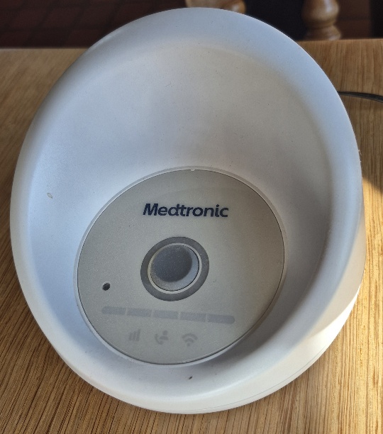 A close-up view of a white, bowl-shaped Medtronic CareLink monitor sitting on a light wooden table surface. The device features a circular center faceplate displaying the Medtronic logo, a central power button, and indicator icons for cellular signal, status, and Wi-Fi connection.