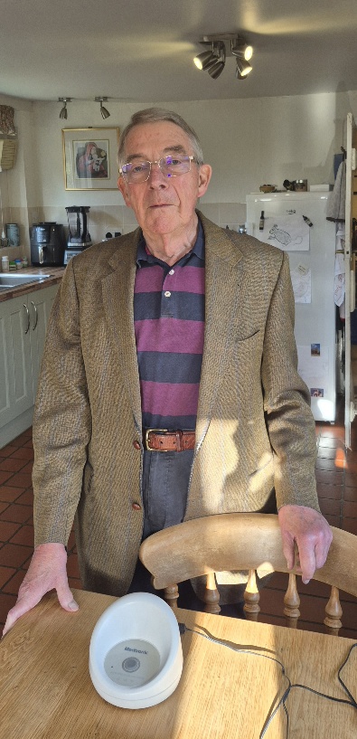 Philip Pelly standing in his kitchen behind a wooden table, resting his hands on the table and a wooden chair back. He wears glasses, a purple and dark-striped polo shirt, and a brown patterned blazer. On the wooden table in front of him rests a small white Medtronic remote monitoring device with a connected power cable.