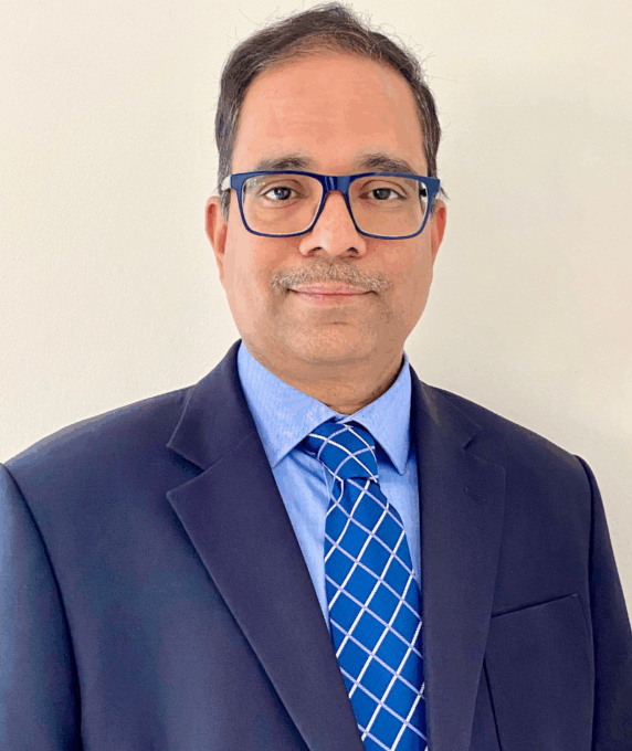 Photograph of interim medical director, Dr Kallur Suresh. He is wearing a navy blue suite jacket, light blue shirt and a diamond patterned blue tie. He is also wearing blue rimmed glasses.