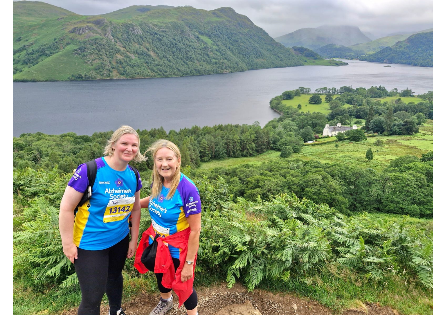 Two women in bright sports wear standing among bushes with a river and mountains behind them.