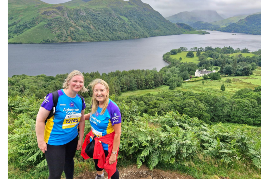 Two women in bright sports wear standing among bushes with a river and mountains behind them.