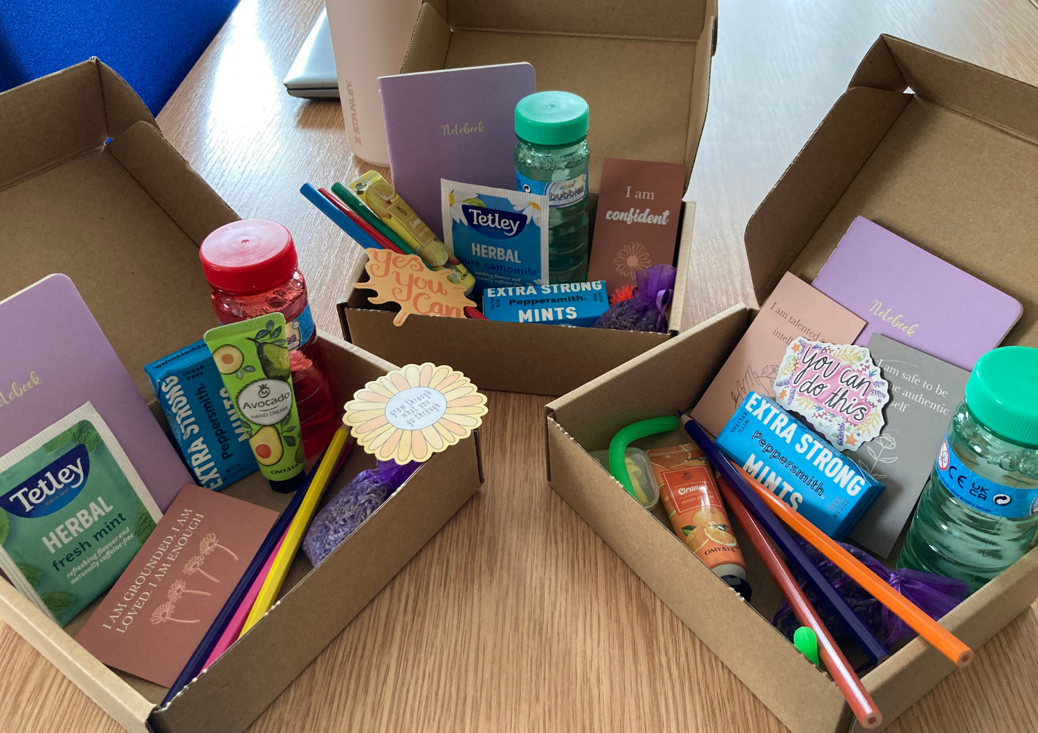 Three open cardboard gift boxes on a wooden table, each filled with care package items including notebooks, herbal tea, mints, colored pencils, hand cream, and affirmative quote cards.