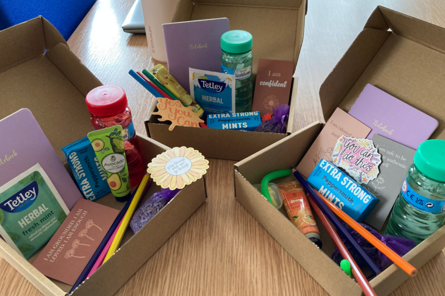 Three open cardboard gift boxes on a wooden table, each filled with care package items including notebooks, herbal tea, mints, colored pencils, hand cream, and affirmative quote cards.