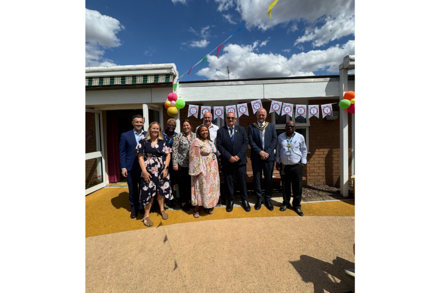 A group of eight people posing outside a building under a banner that reads "GARDEN PARTY" decorated with colorful bunting and balloons.