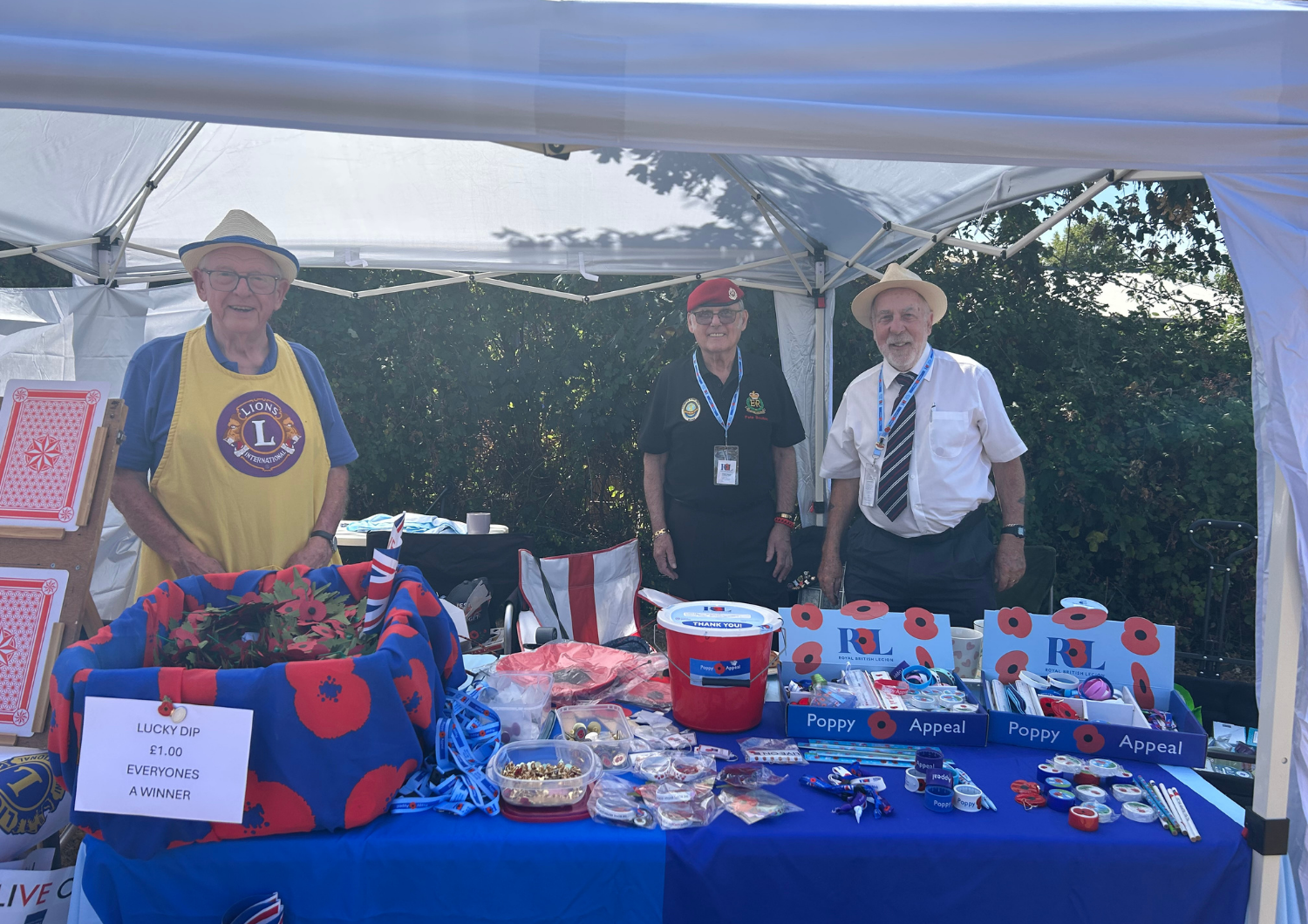Three elderly men standing behind a charity stall under a white gazebo. The table is covered in a blue cloth and filled with Royal British Legion Poppy Appeal items, a red donation bucket, and a 