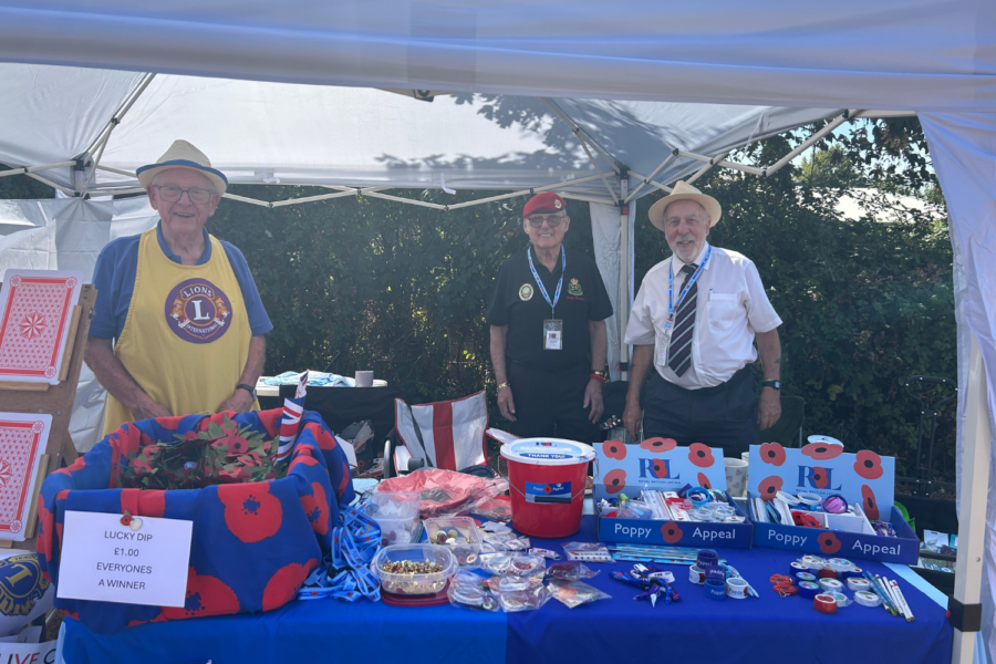 Three elderly men standing behind a charity stall under a white gazebo. The table is covered in a blue cloth and filled with Royal British Legion Poppy Appeal items, a red donation bucket, and a "Lucky Dip" basket.