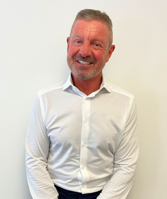 Headshot of Trevor, a smiling man with short hair and a beard, wearing a white long-sleeved button-down shirt against a white backdrop.