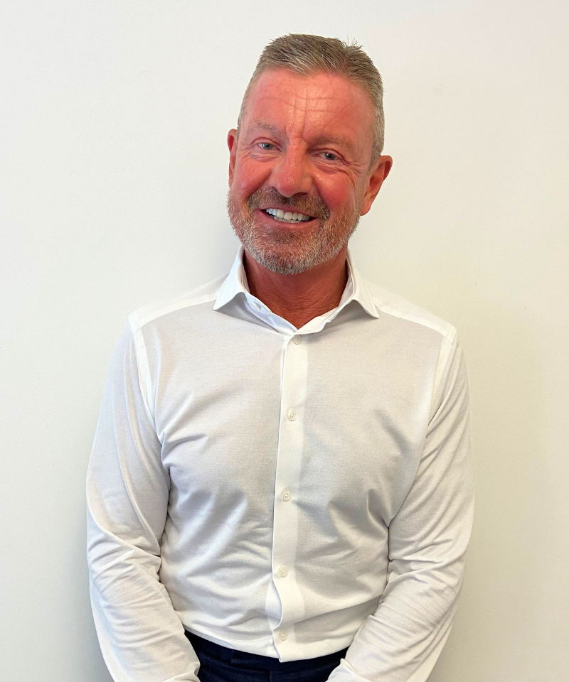 Headshot of Trevor, a smiling man with short hair and a beard, wearing a white long-sleeved button-down shirt against a white backdrop.