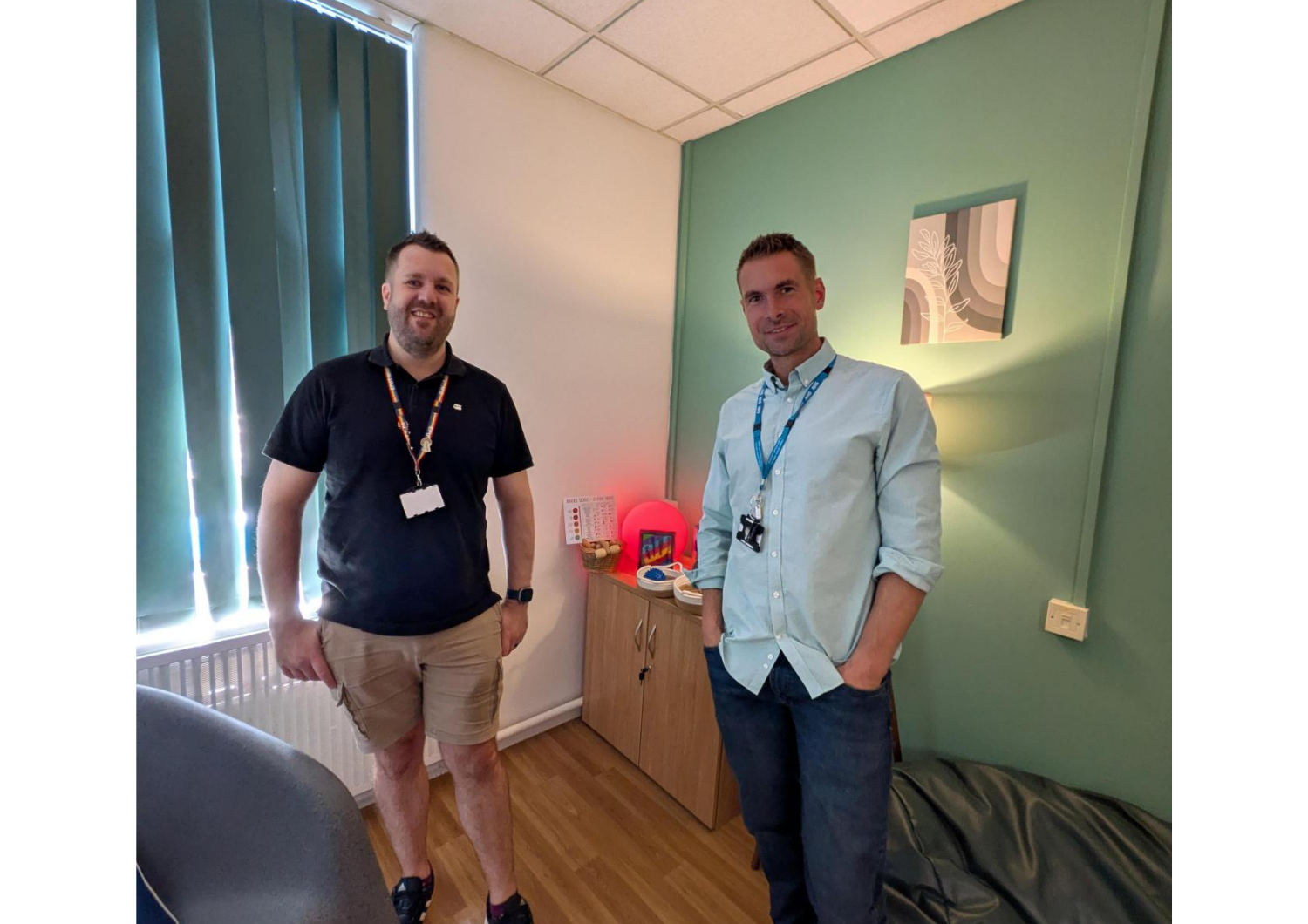 Two NHS healthcare staff members standing inside a calm, newly decorated sensory room with soft lighting, a sage green accent wall, and sensory equipment on a side cabinet.