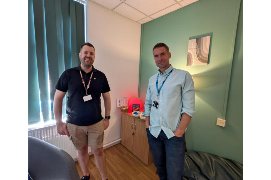 Two NHS healthcare staff members standing inside a calm, newly decorated sensory room with soft lighting, a sage green accent wall, and sensory equipment on a side cabinet.