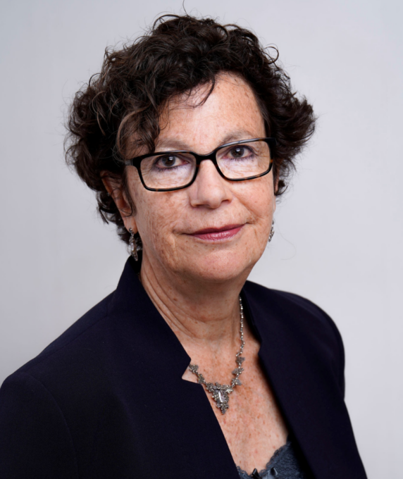Headshot of Jackie Carissati wearing black-rimmed glasses, a dark blazer, and a detailed metallic necklace against a plain off-white background.
