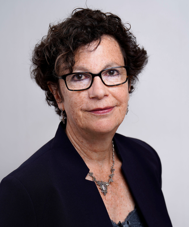 Headshot of Jackie Carissati wearing black-rimmed glasses, a dark blazer, and a detailed metallic necklace against a plain off-white background.