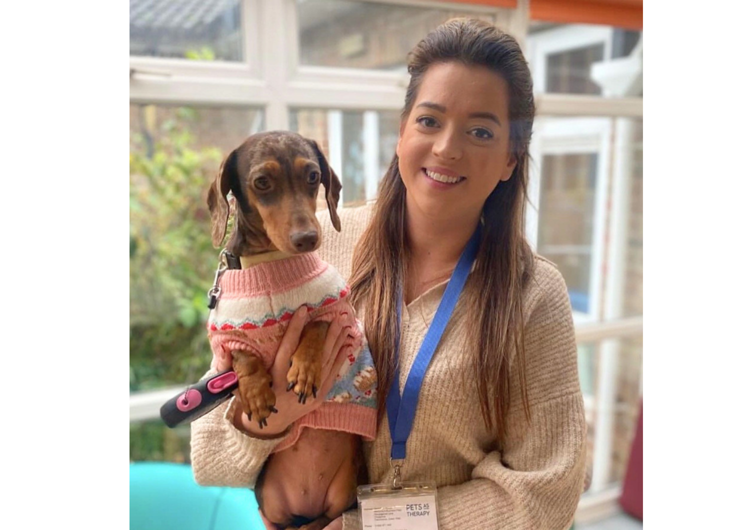 Sylvie smiling while holding Ripple, a dachshund therapy dog wearing a pink patterned jumper.