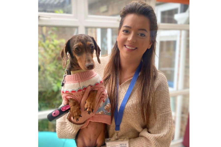 Sylvie smiling while holding Ripple, a dachshund therapy dog wearing a pink patterned jumper.