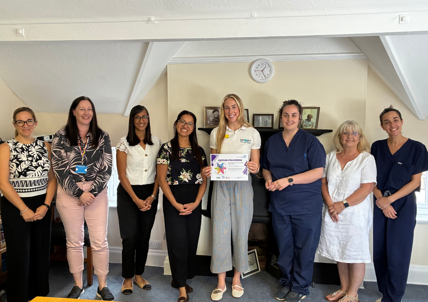 Eight staff members from Tile House and Sport for Confidence pose together indoors while a woman in the center holds up a Certificate of Accreditation.