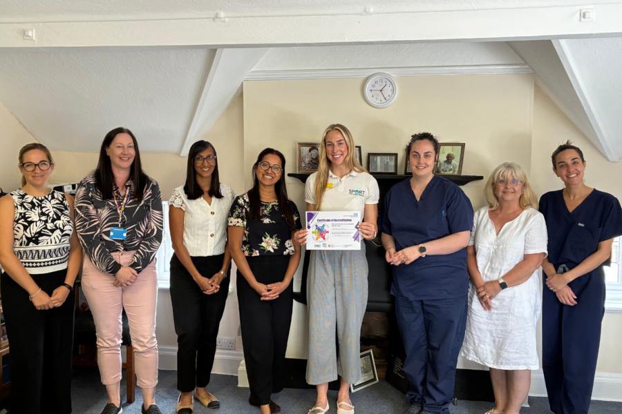 Eight staff members from Tile House and Sport for Confidence pose together indoors while a woman in the center holds up a Certificate of Accreditation.