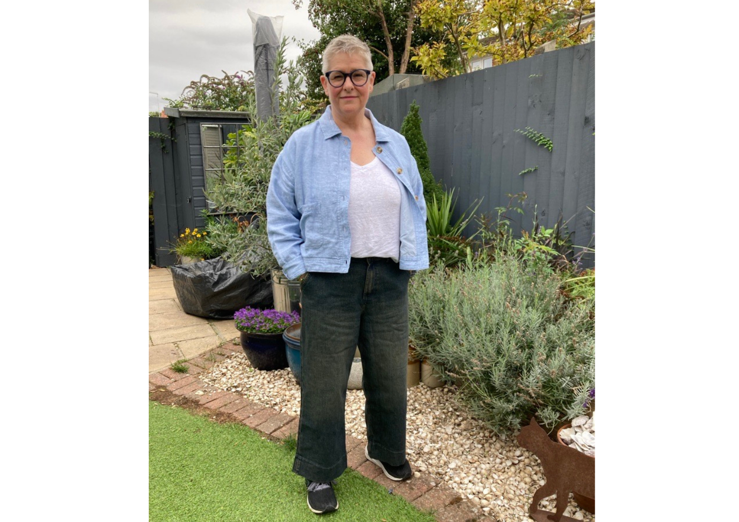 A person with short silver hair and round glasses standing in a garden, wearing a light blue jacket, white top, and dark wide-leg trousers.