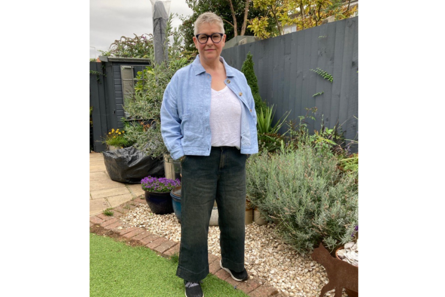 A person with short silver hair and round glasses standing in a garden, wearing a light blue jacket, white top, and dark wide-leg trousers.