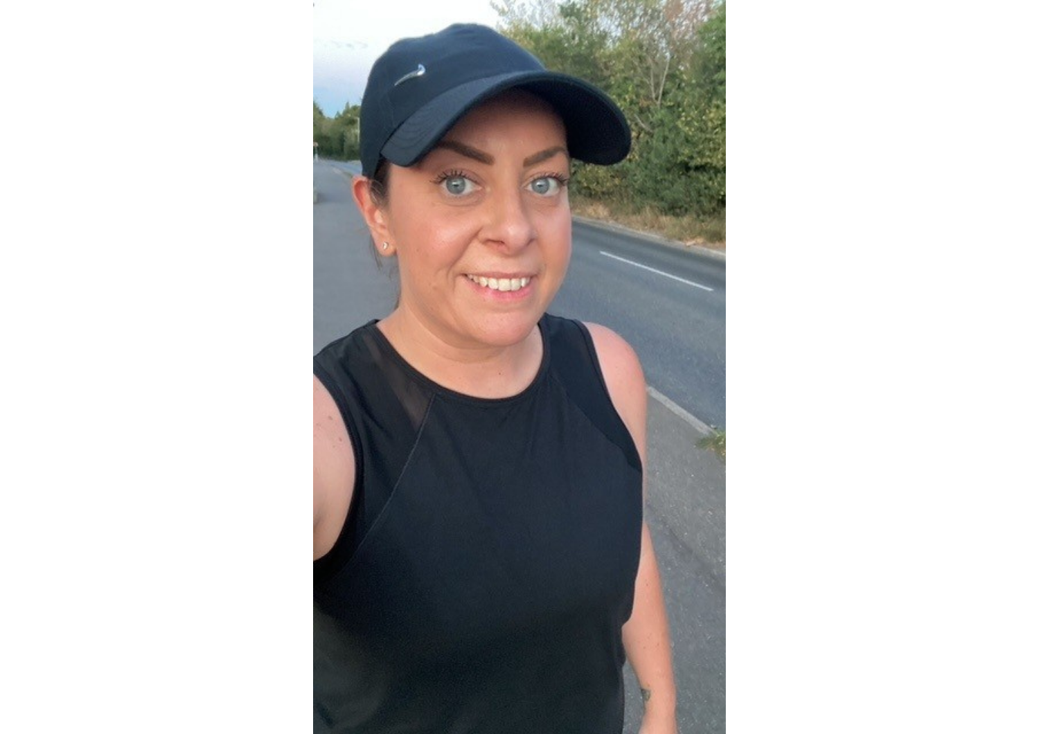 A selfie of a smiling woman outdoors on a pavement alongside a road, dressed in athletic wear including a black sleeveless top and a black Nike baseball cap.