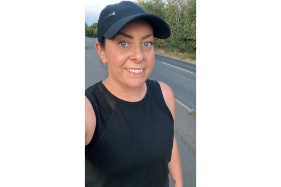 A selfie of a smiling woman outdoors on a pavement alongside a road, dressed in athletic wear including a black sleeveless top and a black Nike baseball cap.
