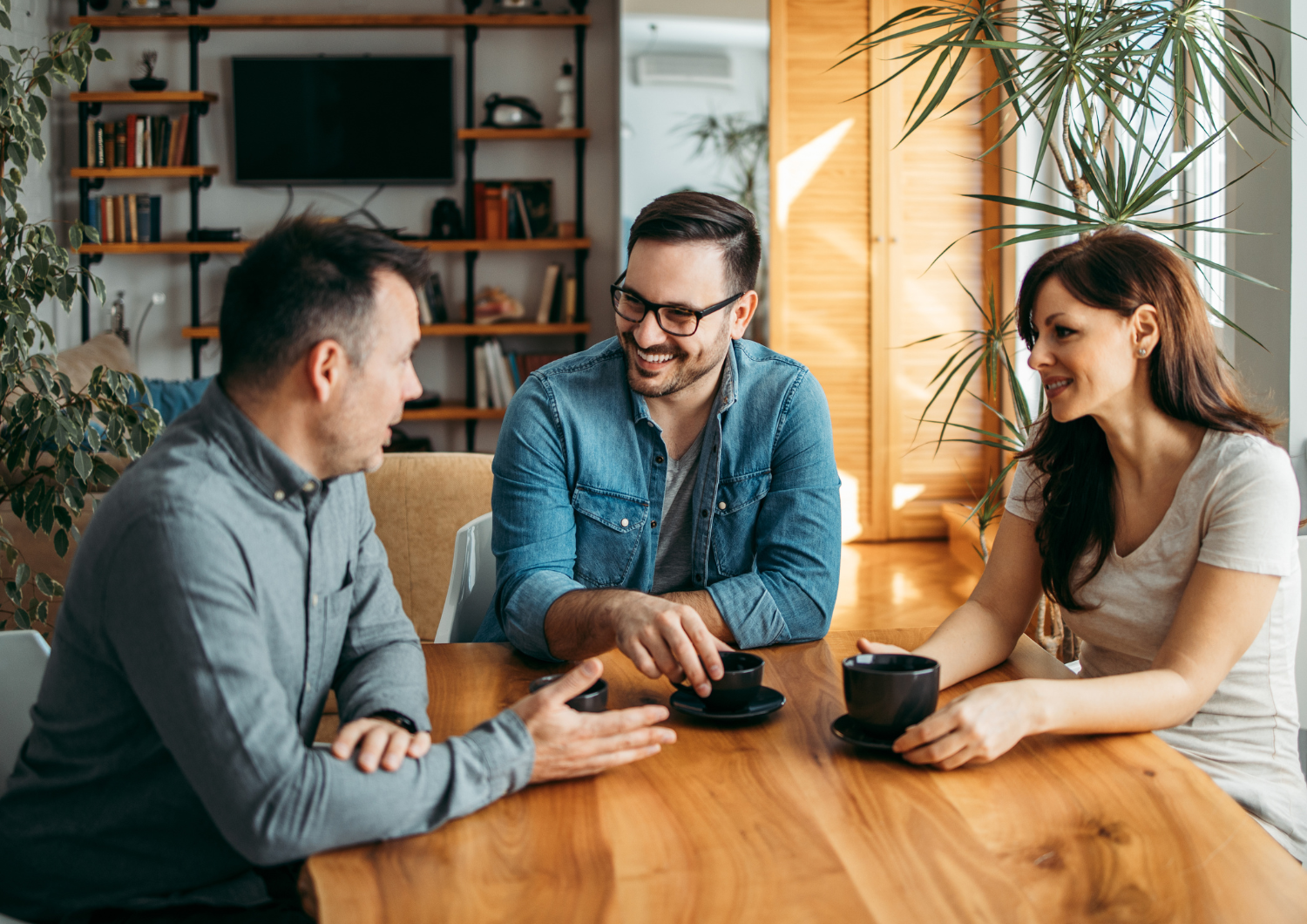 Three adults—two men and one woman—sitting around a wooden table in a brightly lit room with houseplants and bookshelves in the background, having a casual discussion over cups of coffee.