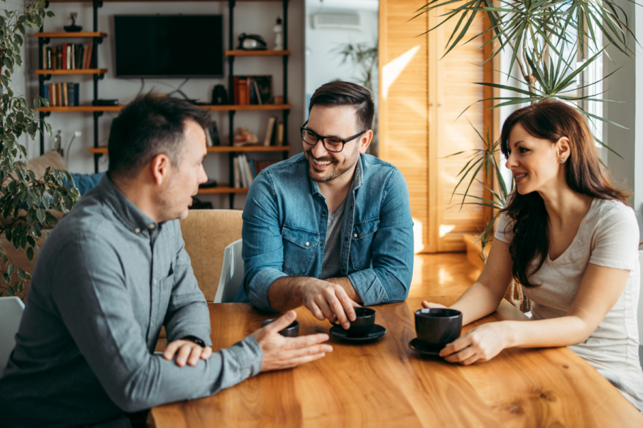 Three adults—two men and one woman—sitting around a wooden table in a brightly lit room with houseplants and bookshelves in the background, having a casual discussion over cups of coffee.