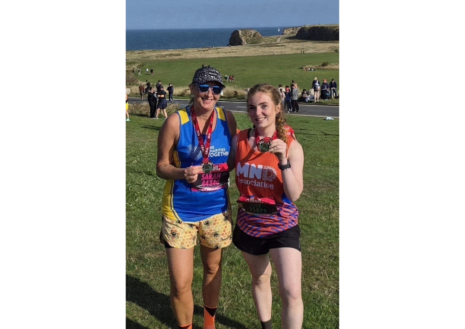 Two women, Sarah and Lilly Parker, standing side by side outdoors on a sunny day after completing a race, both wearing medals around their necks and race bibs on their shirts. The woman on the left wears a blue and yellow vest, a patterned cap, sunglasses, and patterned shorts. The woman on the right wears an orange top with 