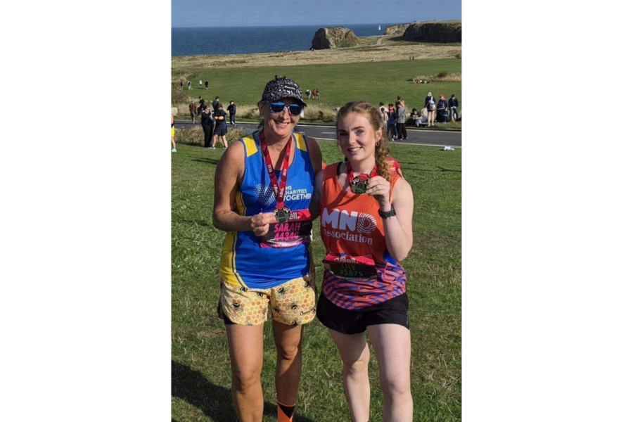 Two women, Sarah and Lilly Parker, standing side by side outdoors on a sunny day after completing a race, both wearing medals around their necks and race bibs on their shirts. The woman on the left wears a blue and yellow vest, a patterned cap, sunglasses, and patterned shorts. The woman on the right wears an orange top with "MND Association" text, dark shorts, and holds up her medal. They are posing in a grassy area with a coastal view, cliffs, and the sea in the background.