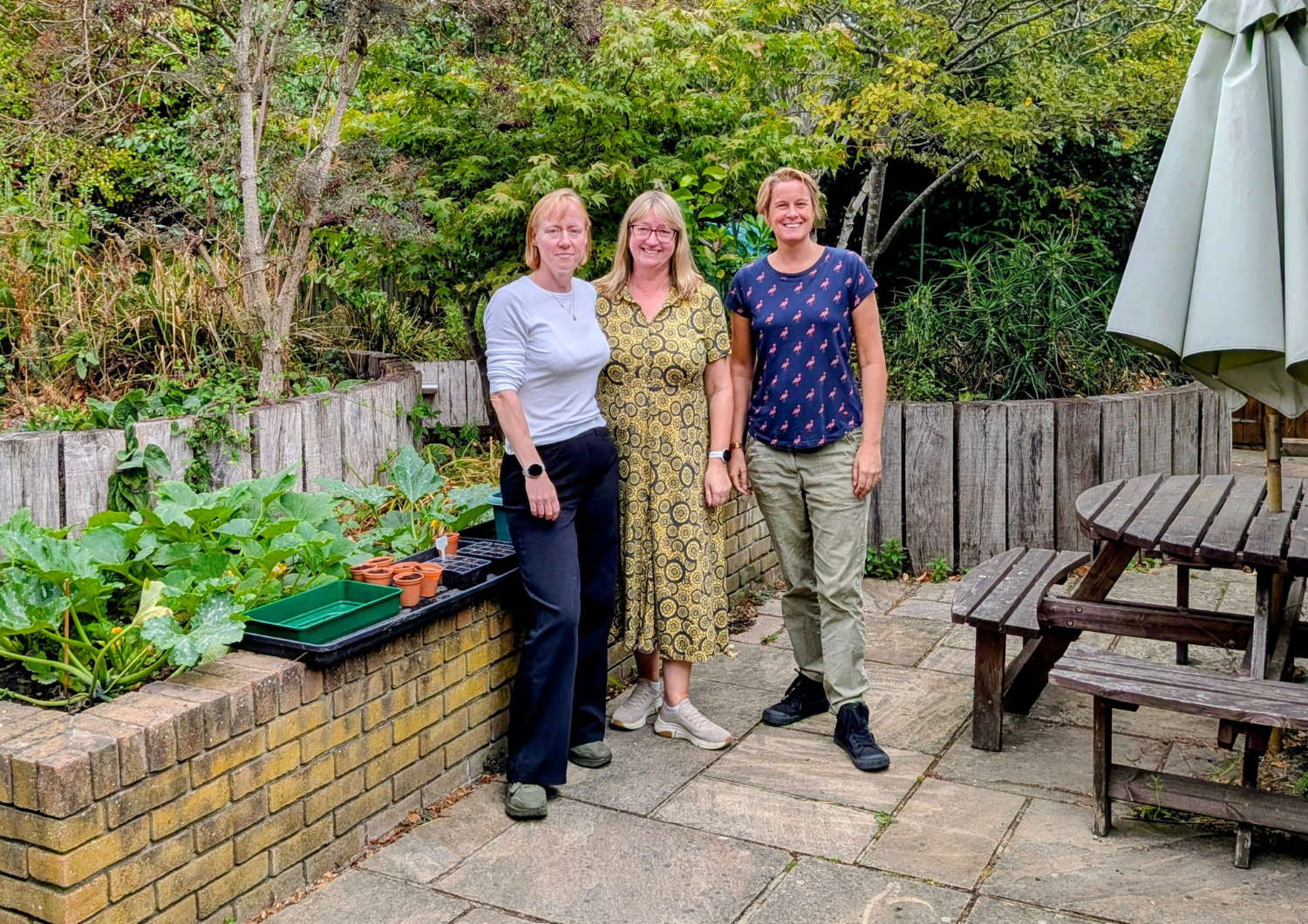 Occupational Therapy team, Kate Jackson, Angela Higgins, Kelly Burgess, standing together and smiling in an outdoor garden patio. They are posed next to a brick raised garden bed with plants and terracotta pots, with a wooden picnic table and patio umbrella on the right.