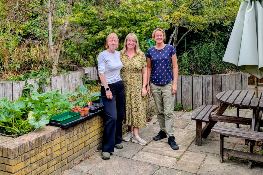 Occupational Therapy team, Kate Jackson, Angela Higgins, Kelly Burgess, standing together and smiling in an outdoor garden patio. They are posed next to a brick raised garden bed with plants and terracotta pots, with a wooden picnic table and patio umbrella on the right.