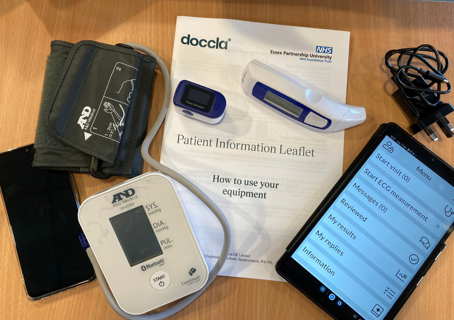 A collection of home health monitoring equipment laid out on a wooden table, including a digital blood pressure monitor with cuff, a blue pulse oximeter, a digital forehead thermometer, charging cables, a smartphone displaying a healthcare app menu, and a Doccla and NHS Essex Partnership University Patient Information Leaflet titled 