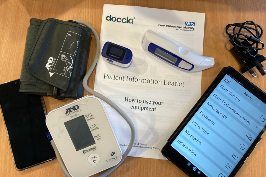 A collection of home health monitoring equipment laid out on a wooden table, including a digital blood pressure monitor with cuff, a blue pulse oximeter, a digital forehead thermometer, charging cables, a smartphone displaying a healthcare app menu, and a Doccla and NHS Essex Partnership University Patient Information Leaflet titled "How to use your equipment".