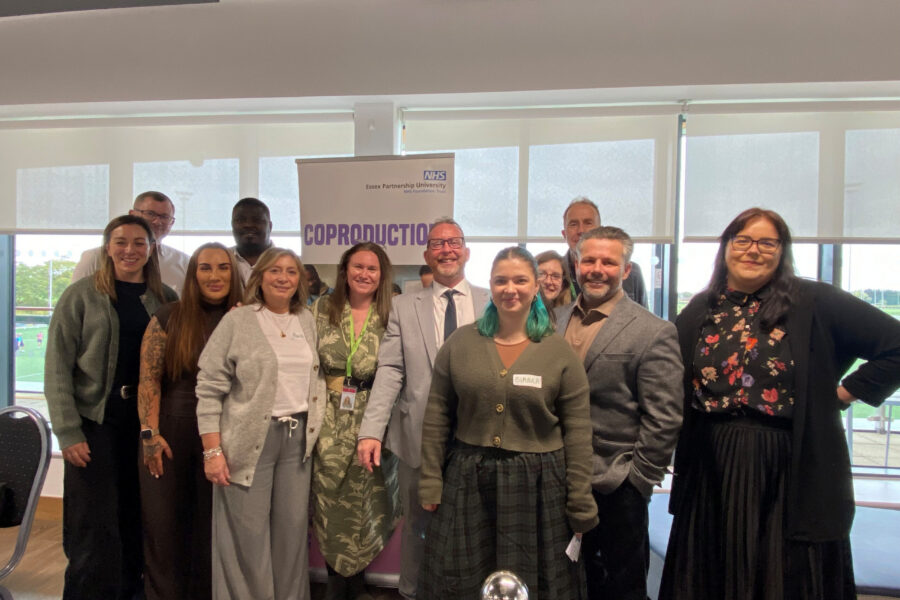A group photo of eleven attendees standing together in front of a banner that reads "NHS Essex Partnership University" and "COPRODUCTION". The group poses indoors with large windows in the background.