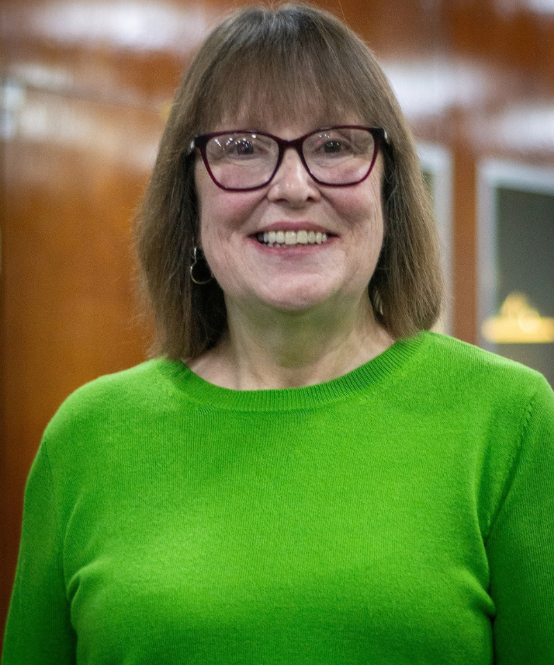 A headshot of a smiling woman with straight shoulder-length brown hair with bangs, wearing glasses with reddish frames and a vibrant bright green crewneck sweater.