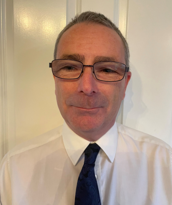 A headshot of a man with short graying hair and glasses, wearing a white collared shirt and a dark blue pattern tie against a light door background.