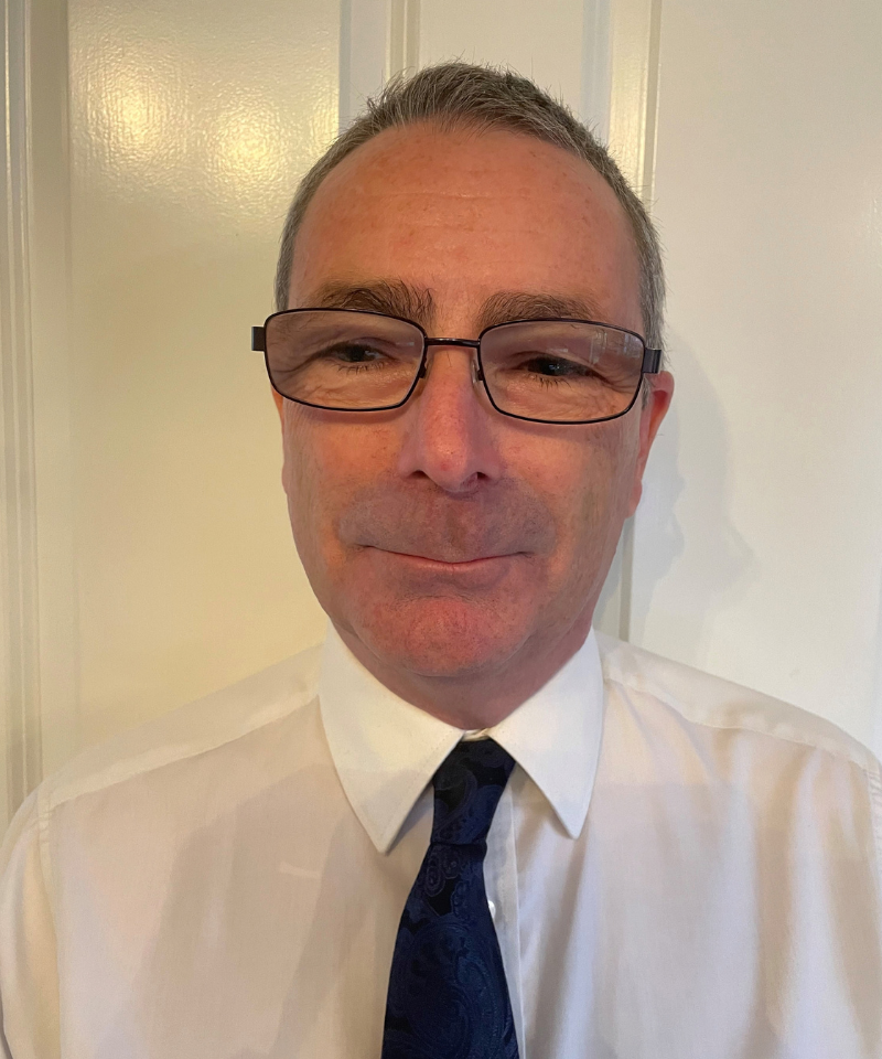 A headshot of a man with short graying hair and glasses, wearing a white collared shirt and a dark blue pattern tie against a light door background.
